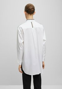 Bílá košile s dlouhým rukávem a zakřiveným lemem, s černým lemem kolem límce a s vertikálním švem na zadní straně. Vyrobena z hladkého materiálu.