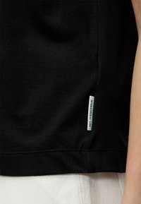 Vêtement noir avec une petite étiquette en tissu blanc près de l'ourlet indiquant « EST. IN STOCKHOLM », porté au-dessus d'un pantalon blanc et un bras visible.