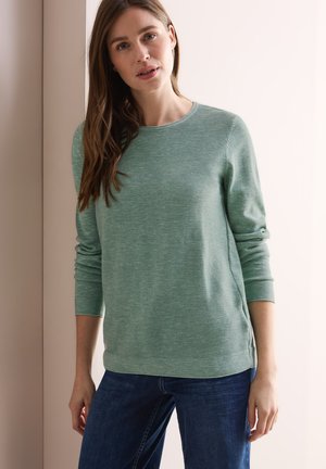 Grüner Langarmpullover mit strukturiertem, geripptem Muster; Rundhals-Ausschnitt und seitlichen Schlitzen; kombiniert mit dunkelblauen Jeans.