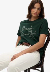 Grøn bomulds T-shirt med et trykt design af "Pinnacles National Park" i lysegrå, parret med hvide jeans og et flettet brunt bælte.