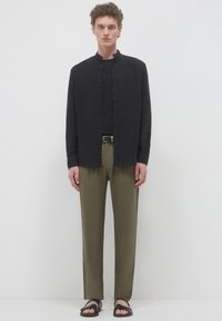 Modello maschile che indossa una camicia nera con colletto alla mandarino, una t-shirt nera, pantaloni verdi oliva con cintura e sandali neri su uno sfondo bianco semplice.