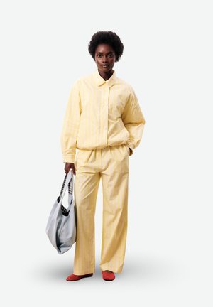 Vrouw draagt een losse geel gestreepte blouse en broek met rode schoenen, en houdt een grote grijze boodschappentas in één hand vast.