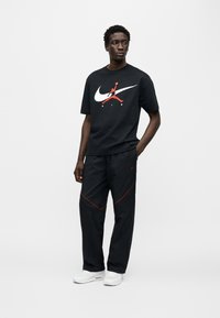 Μαύρο μπλουζάκι Nike με μεγάλο λευκό και κόκκινο λογότυπο, σε συνδυασμό με μαύρο παντελόνι με κόκκινες λεπτομέρειες και λευκά αθλητικά παπούτσια.