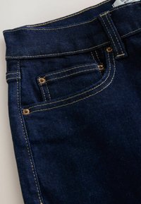 Tamsiai mėlyni džinsai su prigludusia juosta, kontrastinga auksine siūle, ir priekyje su kišene, kuri turi metalines nitų detales.