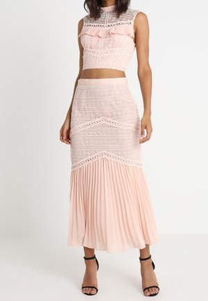 Femme portant un crop top en dentelle sans manches rose pâle et une jupe taille haute assortie avec un ourlet plissé, associée à des talons noirs à bride cheville.