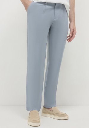 Homme portant un pantalon droit bleu clair et des chaussures beige à enfiler, se tenant devant un fond uni.