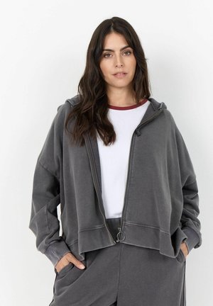 Donna con lunghi capelli scuri che indossa una felpa grigia con zip e pantaloni abbinati sopra una camicia bianca con colletto amaranto, in piedi con le mani nelle tasche.