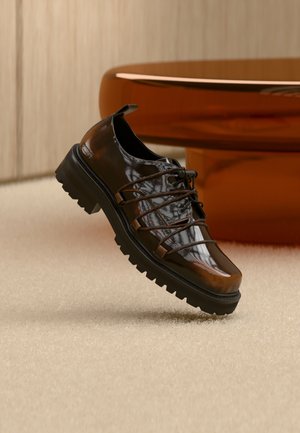 Glossy brown leather shoe with thick black sole and unique lacing, positioned on beige carpet near rounded orange furniture.