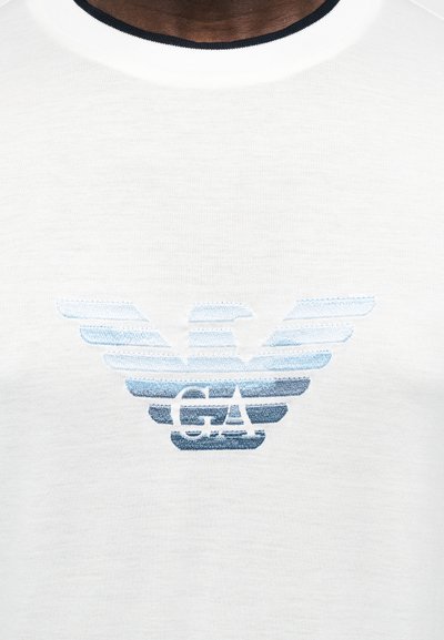 Κοντινή λήψη λευκού πουκαμίσου με κεντητό, ανοιχτό μπλε λογότυπο αετού GA στο κέντρο του στήθους.