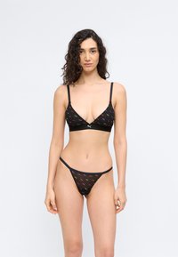 Γυναίκα με σγουρά μαλλιά που φοράει μαύρο σουτιέν και τάνγκα σετ με πολύχρωμα λογότυπα Puma σε λευκό φόντο.