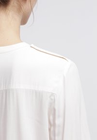 Blouse blanche à la texture lisse et soyeuse, avec un empiècement dos et un petit détail de bouton sur l'épaule pour un design fonctionnel.