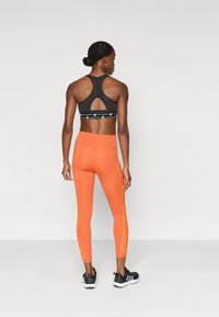 Μαύρο αθλητικό σουτιέν με άνοιγμα στην πλάτη, σε συνδυασμό με πορτοκαλί εφαρμοστά κολάν που έχουν μαύρες ρίγες στο πλάι και ραφές λεπτομερειών.