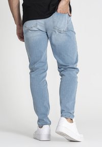 Vaqueros de mezclilla azul claro con un ajuste slim, piernas ajustadas y dos bolsillos traseros. Llevados con zapatillas blancas, con una textura suave.