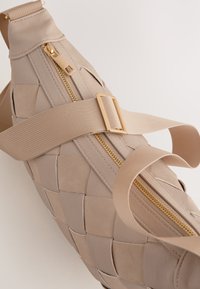 En vävd beige väska med en gyllene dragkedja och tygremmar. Den texturerade ytan har en flätad design med släta detaljer.