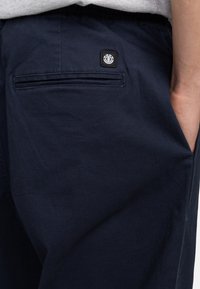 Mørkeblå bukser lavet af glat stof har en elastisk talje, sidelommer og en baglomme med et firkantet logo-patch.