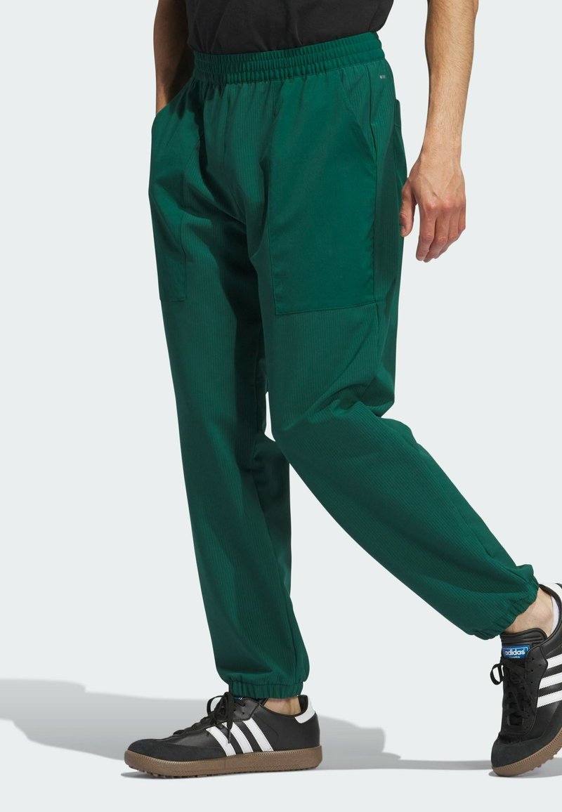 Grüne, elastische Bundhosen mit lockerer Passform. Verfügt über Seitentaschen und einen leichten, strukturierten Stoff. Kombiniert mit schwarzen Schuhen.