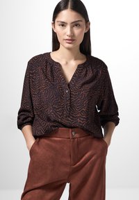 Schwarz und rostfarben gemustertes Hemd mit V-Ausschnitt und Knopfdetails, kombiniert mit rustfarbenen, glatt strukturierten, hoch-taillierten Hosen.