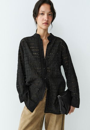 Junge Frau trägt eine schwarze, durchsichtige Bluse mit Knöpfen und langen Ärmeln, beige Hose, hält eine schwarze Clutch vor einfarbigem Hintergrund.