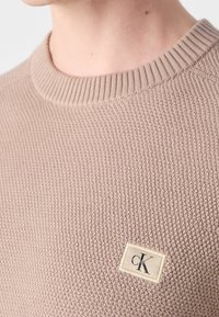 Gros plan sur un pull en maille texturée beige avec un col rond côtelé et une petite étiquette rectangulaire du logo Calvin Klein sur la poitrine.