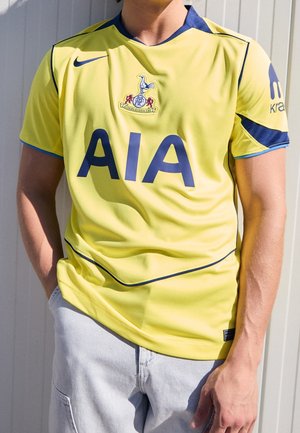 Osoba ubrana w żółtą koszulkę piłkarską Tottenham Hotspur z ciemnoniebieskim obszyciem, dużym napisem "AIA" oraz widocznymi logo sportowymi.
