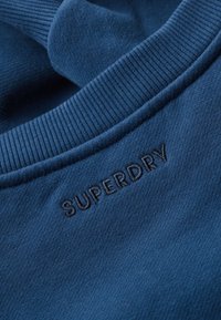 Zbliżenie niebieskiego dzianego ubrania z prążkowanym kołnierzem i wyhaftowanym logo „SUPERDRY” w pobliżu dekoltu.