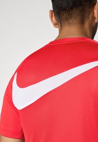 Piros sport póló, amely texturált anyagból készült, és nagyméretű fehér Nike pipa logó díszíti a hátsó vállrészt.