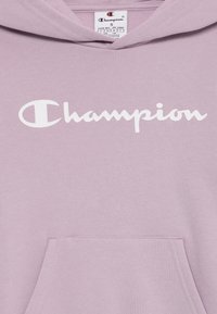 Hoodie violet clair en tissu doux avec une poche avant et le logo "Champion" blanc sur la poitrine.