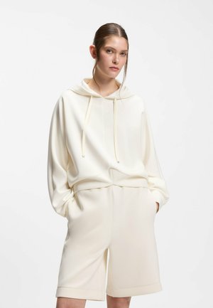 Frau trägt einen cremeweißen Kapuzenpullover mit Kordeln und passende knielange Shorts, steht mit den Händen in den Taschen.