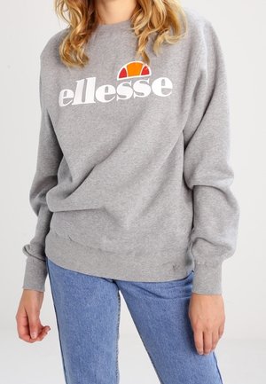 Person trägt ein graues ellesse Sweatshirt mit Logo und blaue Jeans, steht vor einem schlichten hellen Hintergrund.