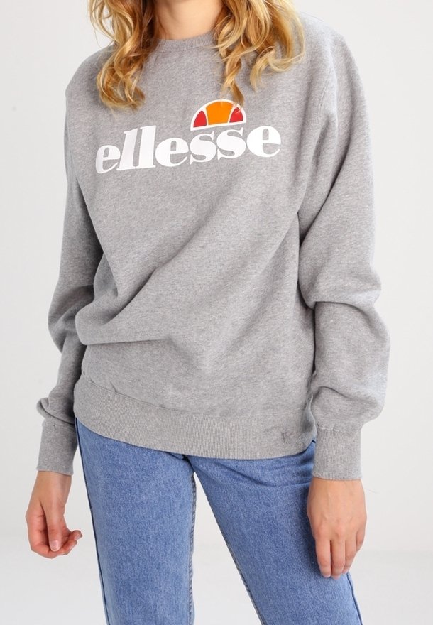 Personne portant un sweat-shirt gris ellesse avec logo et un jean bleu, debout devant un fond clair uni.