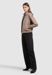 Femme portant une veste beige zippée avec des poches, un haut noir, un jean noir à jambes larges et des bottines à talons noires, debout devant un fond uni.