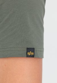 Olivovo zelené bavlnené tričko s krátkymi rukávmi. Obsahuje čiernu etiketu s zlatým logom pri leme. Hladký povrch.
