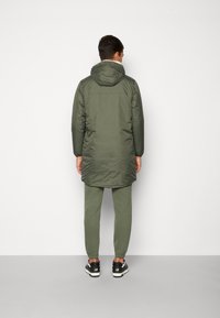 Padded olive green parka with a hood, paired with matching joggers. Features elastic cuffs and side pockets, worn with black athletic shoes.