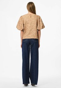 Top beige testurizzato con maniche a sbuffo, dettaglio a occhio di chiave sulla schiena, abbinato a jeans a gamba larga blu scuro a vita alta. Tessuto liscio, stile casual.