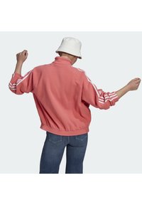Fleece jacket in coral pink with a high collar and white three-stripe design on sleeves. Relaxed fit, elastic hem, and reversible cuffs.