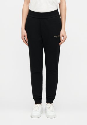 Oseba v črnih hlačah Armani Exchange z elastičnimi manšetami in belimi supergami, stoji pred enobarvnim ozadjem.