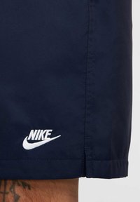 Granatowe szorty wykonane z gładkiego materiału, z białym logo Nike na dolnym lewym rogu. Dolna część jest prosta z delikatnym detalem szwu.