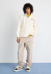 Pullover bianco in pile con collo a zip, abbinato a pantaloni beige e sneakers nere con dettagli bianchi. Il modello si trova su un pavimento blu.