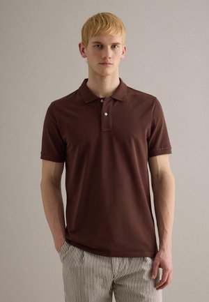 Giovane con capelli biondi corti che indossa una polo marrone a maniche corte e pantaloni a righe, in piedi contro una parete chiara e uniforme.