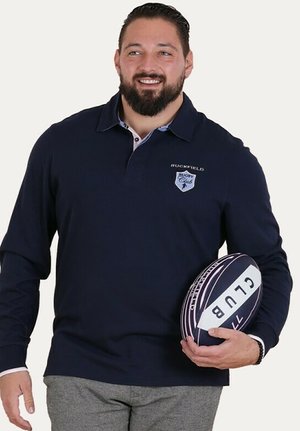 Polo shirt à manches longues bleu marine avec un col contrasté, présentant le logo "Ruckfield" et un ballon de rugby tenu dans la main gauche.