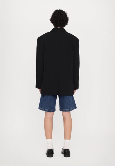 Black oversized blazer made of structured fabric, paired with light blue denim shorts. Black shoes and white socks visible below.