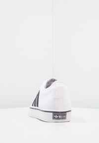 Vita canvas-sneakers med svarta randiga detaljer, platt gummisula och en märkesflik på hälen med texten "Nizza." Lätt och låg profil design.