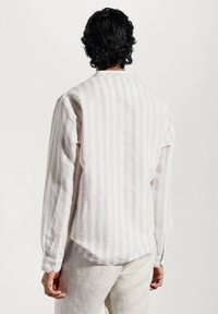 Hombre con cabello rizado oscuro de espaldas, vistiendo una camisa de rayas beige y blanca de manga larga y pantalones de color claro.