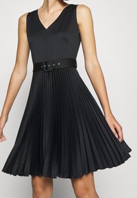 Svart plisserad klänning med V-ringning, ärmlös design och ett skärp i midjan. Har en slät textur och en flödande siluett.