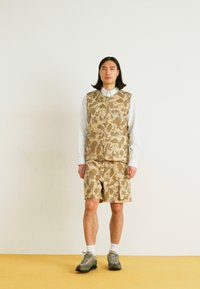 Beige kamouflagedress och shorts-set, gjort av texturerat tyg, med fickor och knappstängning, kombinerad med en vit skjorta med krage.