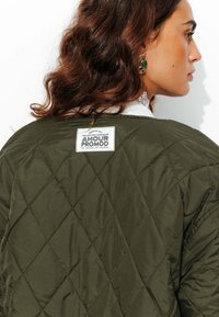 Veste matelassée verte avec un motif en diamant, col blanc et une étiquette portant la mention "Amour Promod" au dos. Tissu texturé.
