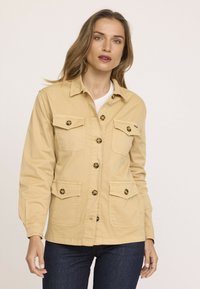 Veste en coton beige avec quatre poches avant et fermeture à boutons. Dotée d'un col classique et de boutons ronds à motifs. Design ajusté.