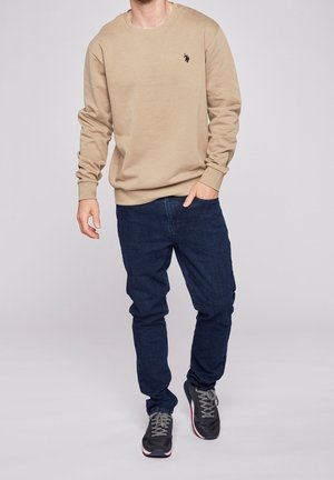 Beżowa bluza z długimi rękawami i okrągłym dekoltem, z małym logo na piersi. Połączona z ciemnoniebieskimi jeansami i szarymi butami sportowymi.