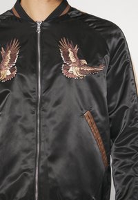 Veste bomber en satin noir avec deux aigles brodés marron sur la poitrine, une fermeture éclair centrale argentée et des poches bordées de marron.
