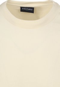 Crème-kleurige katoenen T-shirt met een ronde hals en een effen textuur. Het label vermeldt "SUITABLE" in een donkere lettertype. Geen patronen of accenten zichtbaar.